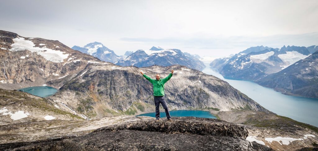 man in green jacket with arms raised standing on a mountain in greenland on an expedtiion cruise