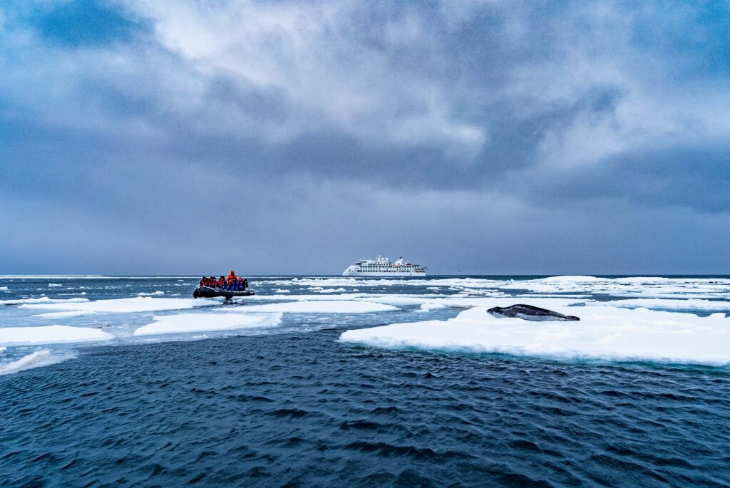 Antarctica travelers sitting in a black zodiac among white icebergs looking at a leopard seal with a small expedition ship in the background