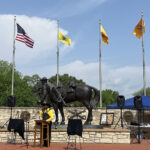 A master of ceremonies stands at a podium in front of a large bronze statue of a Black cavalryman leading his horse, and four flags.