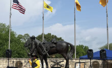 A master of ceremonies stands at a podium in front of a large bronze statue of a Black cavalryman leading his horse, and four flags.