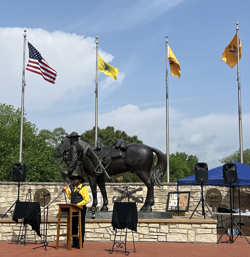 A master of ceremonies stands at a podium in front of a large bronze statue of a Black cavalryman leading his horse, and four flags.